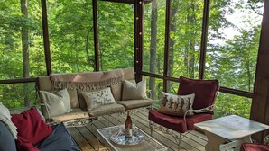 Un área de estar al aire libre cubierta con un sofá, sillas y una mesa, rodeada de amplias ventanas que dan a un bosque.