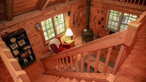 Interior de una cabaña de madera con escalera, chimenea, un sillón rojo y una estantería.
