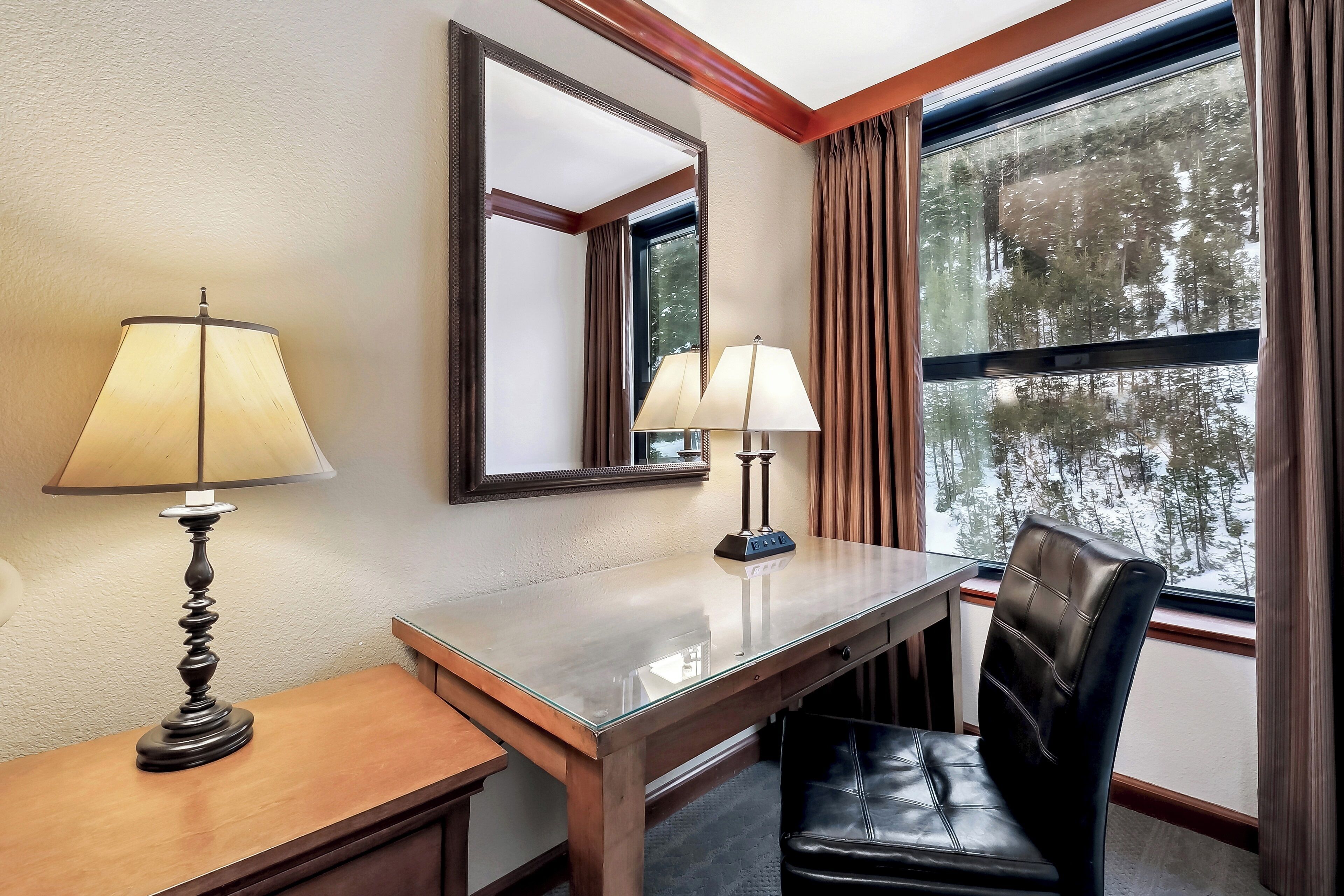 A hotel room with a wooden desk, two lamps, a chair, a large mirror, and a window with curtains.