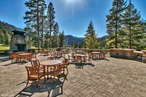 An outdoor patio with wooden tables and chairs, surrounded by tall trees and a stone fireplace. - Located @ Everline Resort & Spa, Pools/Spas/Golf/Ski-in&out/Sledding/Ice Skating (Olympic Valley)