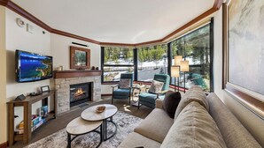 Una sala de estar acogedora con chimenea, asientos cómodos, televisor y ventanas amplias con vistas a un paisaje nevado.