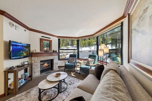 A cozy living room with a fireplace, comfortable seating, a television, and large windows offering a view of a snowy landscape. - Located @ Everline Resort & Spa, Pools/Spas/Golf/Ski-in&out/Sledding/Ice Skating (Olympic Valley)