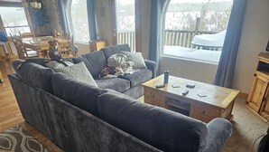 Ein gemütliches Wohnzimmer mit einem grauen Ecksofa, einem Holztisch und Blick auf eine verschneite Terrasse durch große Fenster.