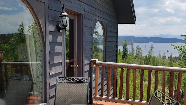 Eine Holzhütte mit Glastisch und Stühlen, einer Laterne und Blick auf einen See und Berge.
