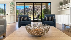 A modern living room with a large glass door leading to a patio with a mountain view. There are two blue sofas with striped cushions, a wooden coffee table, and a kitchen area with white cabinets and a plant shelf.