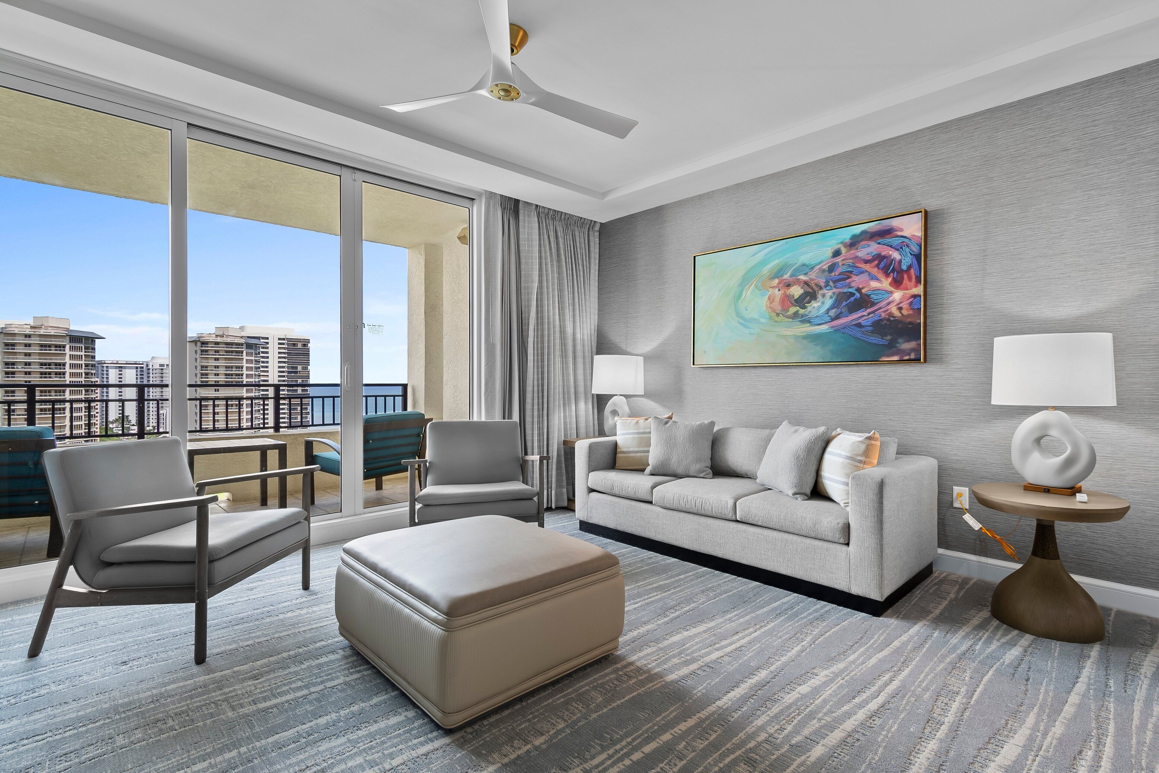 A modern hotel room with a large balcony, a grey sofa, a beige armchair, a round coffee table, and a painting on the wall.