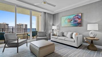 A modern hotel room with a large balcony, a grey sofa, a beige armchair, a round coffee table, and a painting on the wall.