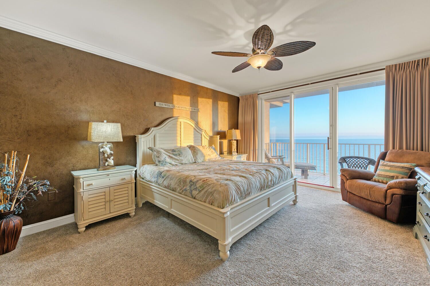 A bedroom with a large bed, a ceiling fan, a nightstand, a chair, and a view of the ocean.