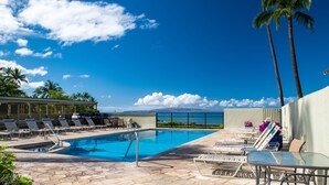 Zona de piscina con sillones de descanso y una mesa, rodeada de palmeras y un cielo despejado.