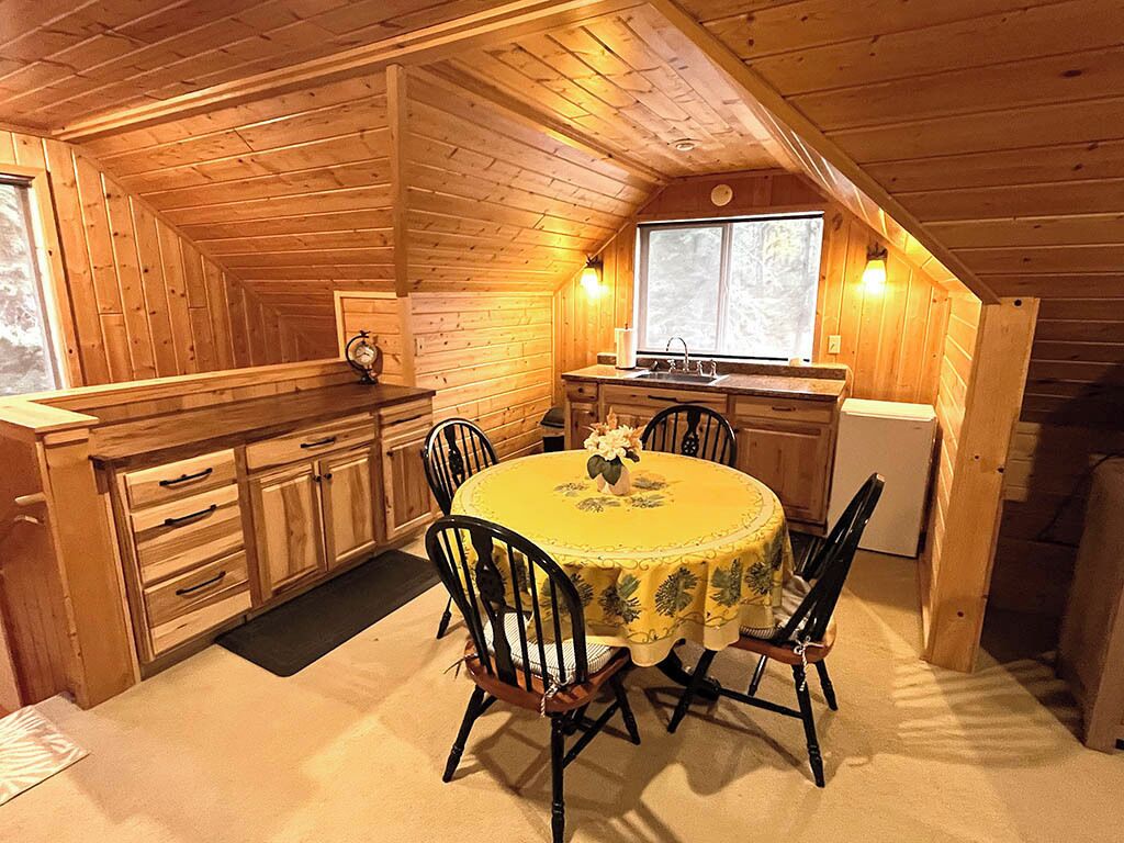 A wooden cabin interior with a dining area featuring a round table and chairs, a kitchen with a window, and wooden walls.