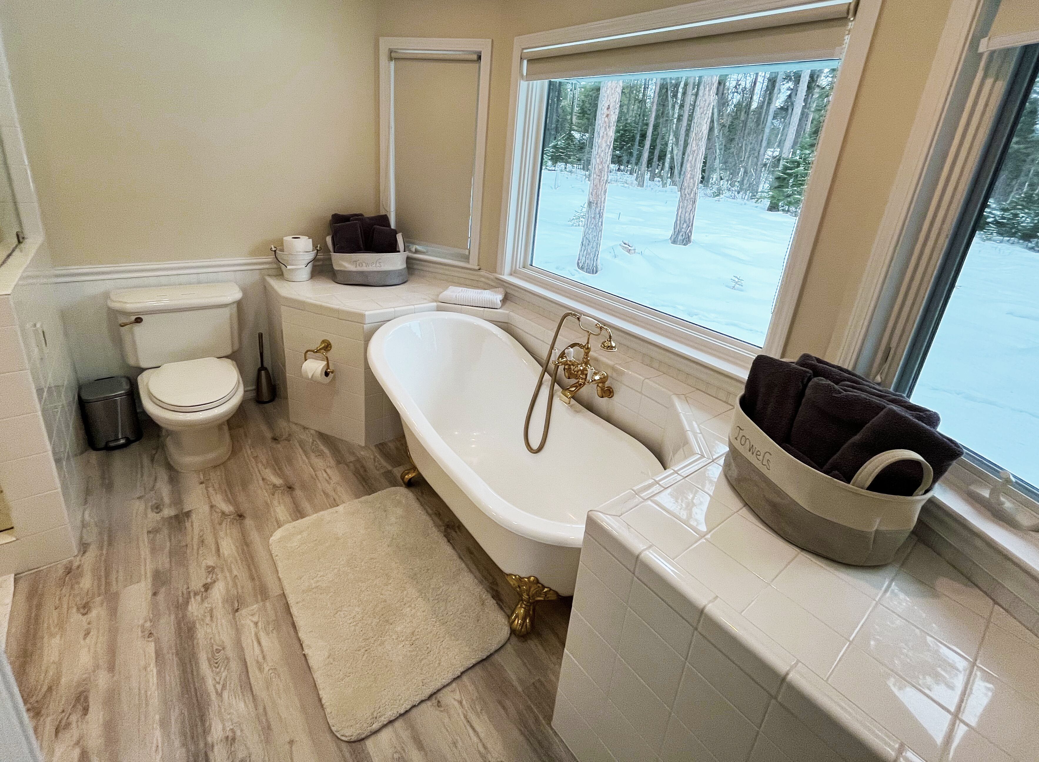 A bathroom with a toilet, a bathtub with a shower, a window with a view of trees, and a towel basket.