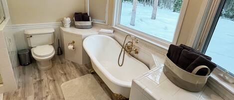 A bathroom with a toilet, a bathtub with a shower, a window with a view of trees, and a towel basket.