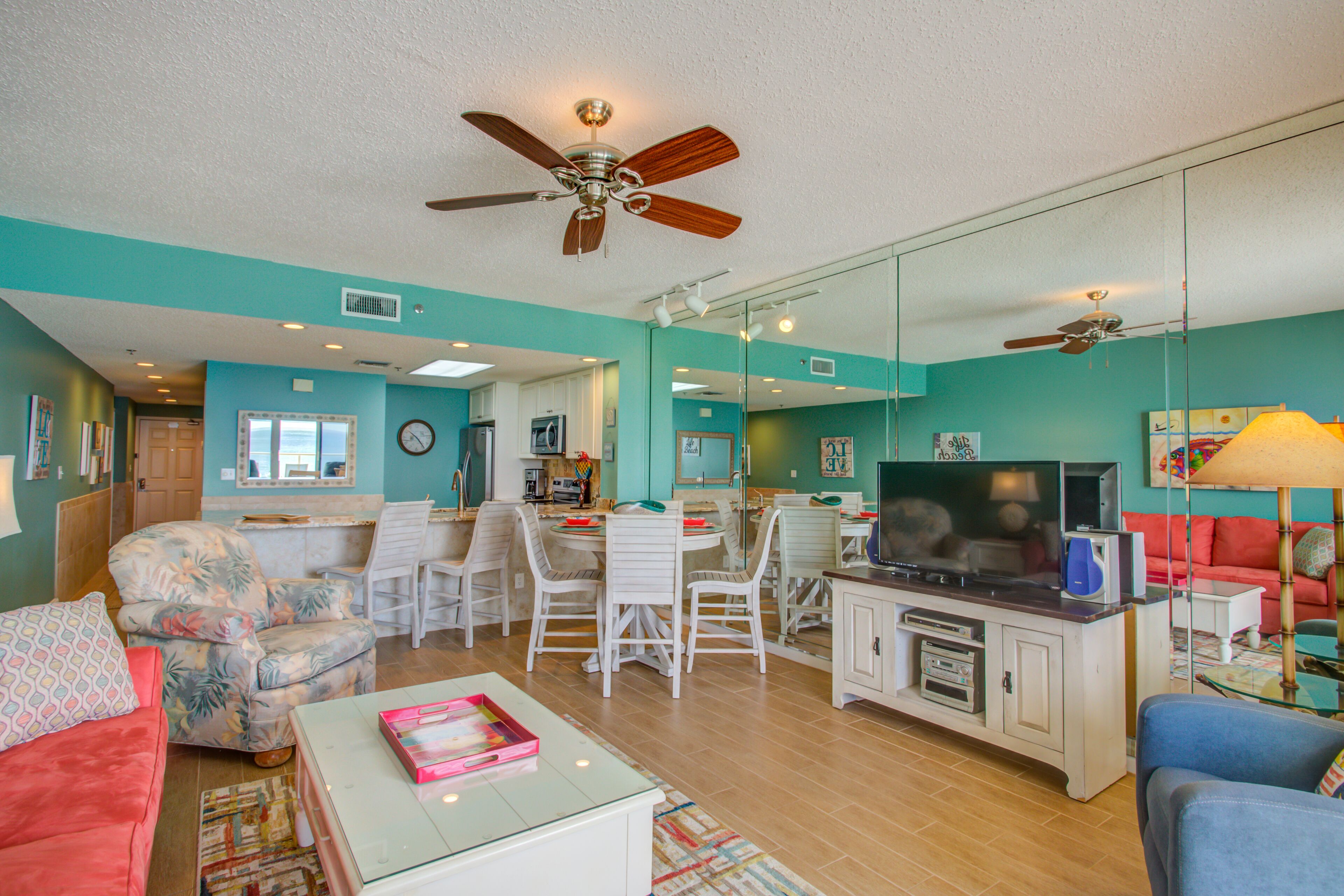 A spacious living room with a ceiling fan, a dining area with chairs and a table, a TV, and a kitchen area with cabinets and appliances.