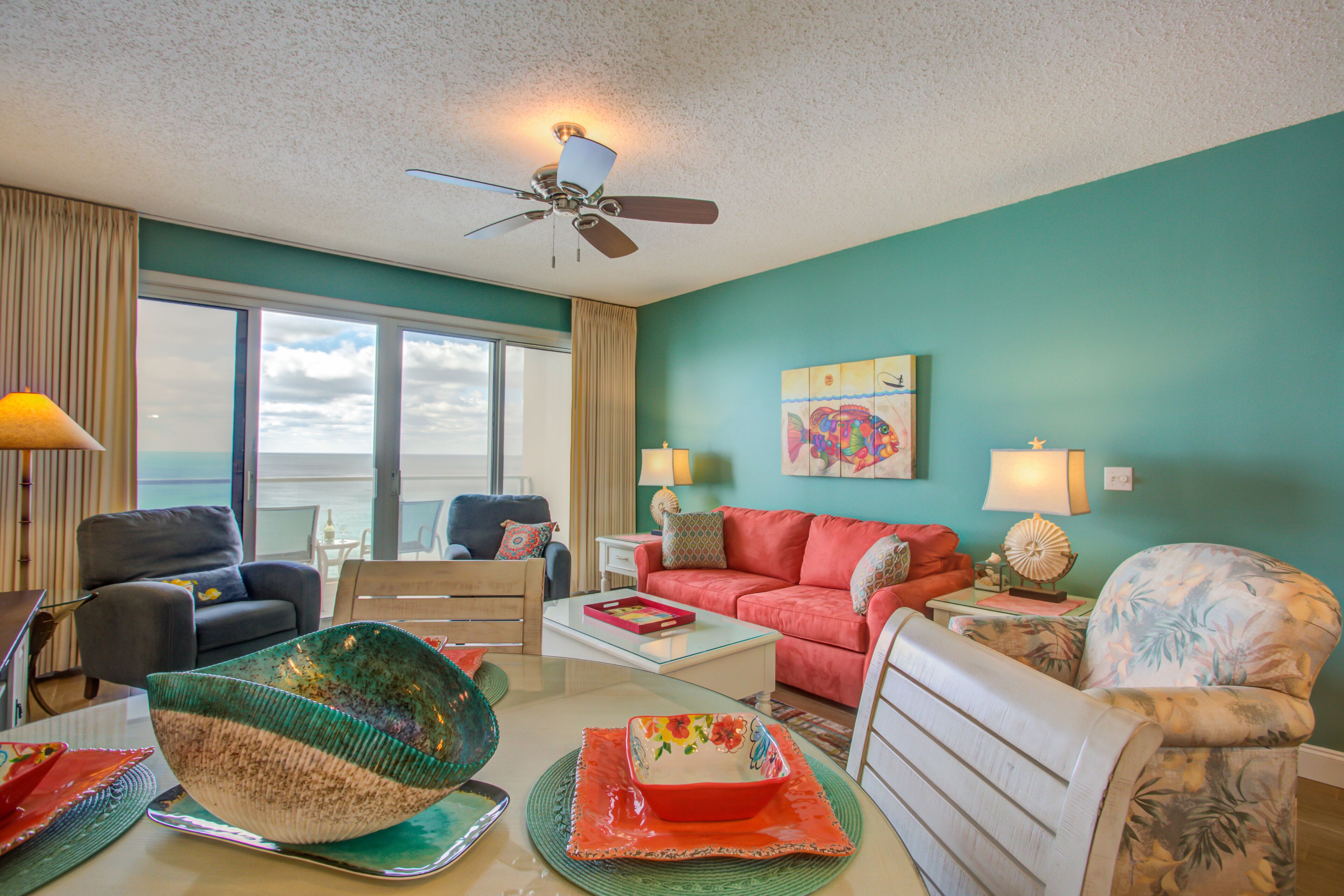 A living room with a view of the ocean, a ceiling fan, and a teal wall.