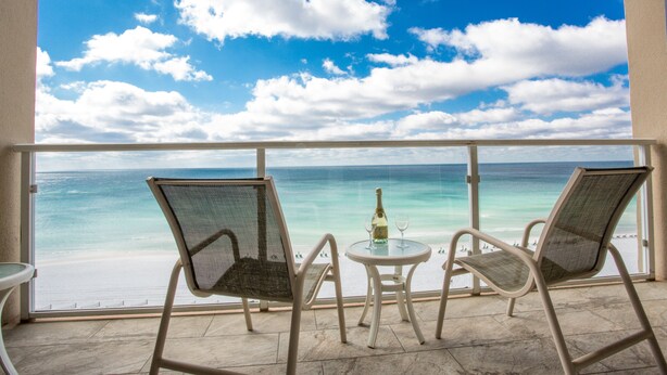 En balkong med två stolar och ett bord, med utsikt över en strand med kristallklart blått vatten och en molnig himmel.