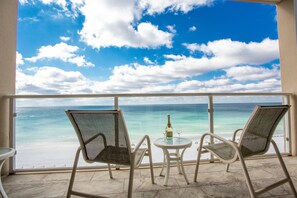 A balcony with two chairs and a table, overlooking a beach with clear blue water and a cloudy sky. - Relax on your balcony with a stunning view of the gulf.   (Destin)