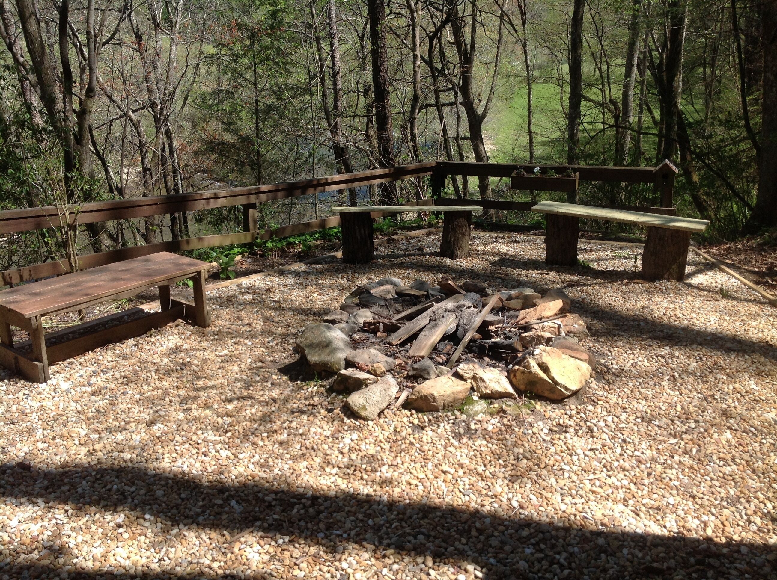 A forested campsite with wooden benches and a fire pit surrounded by gravel.