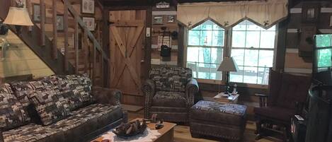 A rustic living room with a wooden staircase, a patterned sofa, a wooden coffee table, and a window with curtains.
