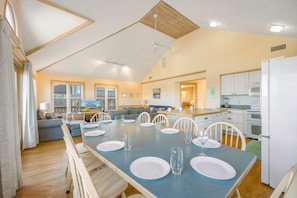 A spacious living area with a dining table set for four, a kitchen with white appliances, and a large window overlooking a stone building. - Spacious Oceanview Home in Frisco with Hot Tub, Screened Porch. Beach Nearby! (Frisco)