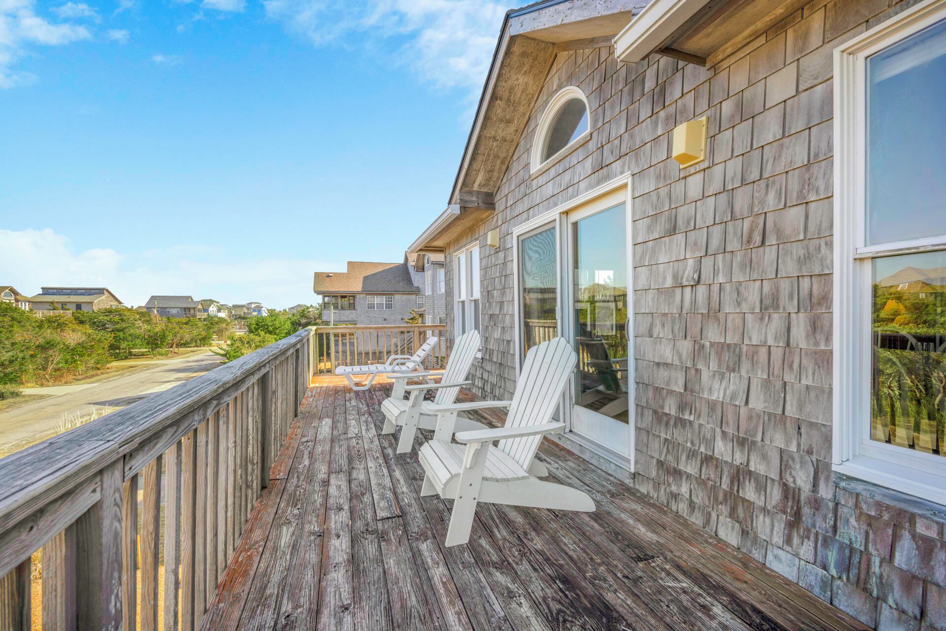 Une terrasse en bois avec deux fauteuils Adirondack blancs, une porte vitrée et une vue sur la plage et les autres maisons.