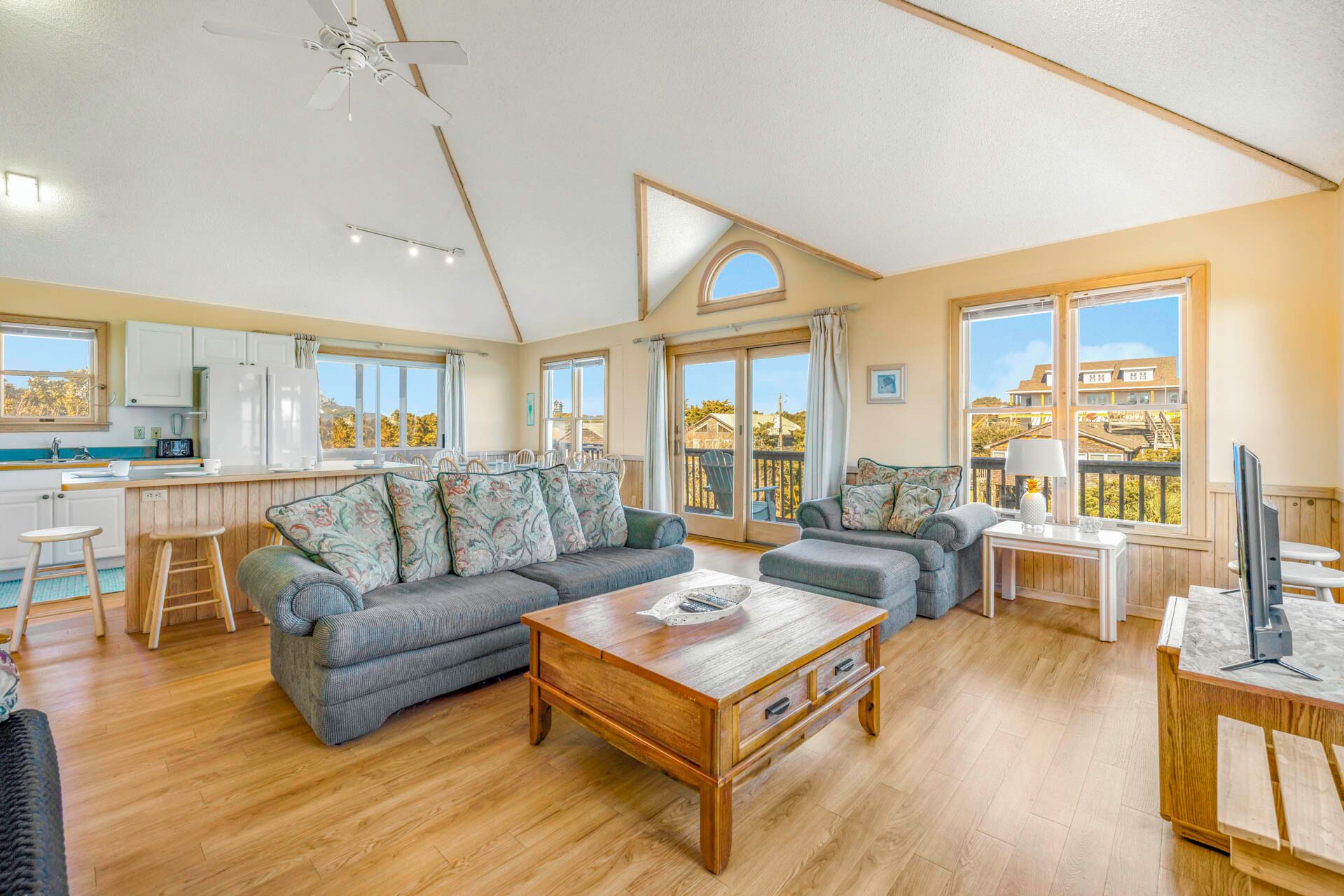 A spacious living area with a sofa, coffee table, and a dining table. There is a kitchen area with cabinets and a window. The room has large windows and a view of the outside.