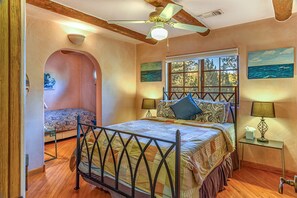 Une chambre à coucher avec un lit, une fenêtre donnant sur un beau paysage, un ventilateur de plafond et une petite table avec une lampe.