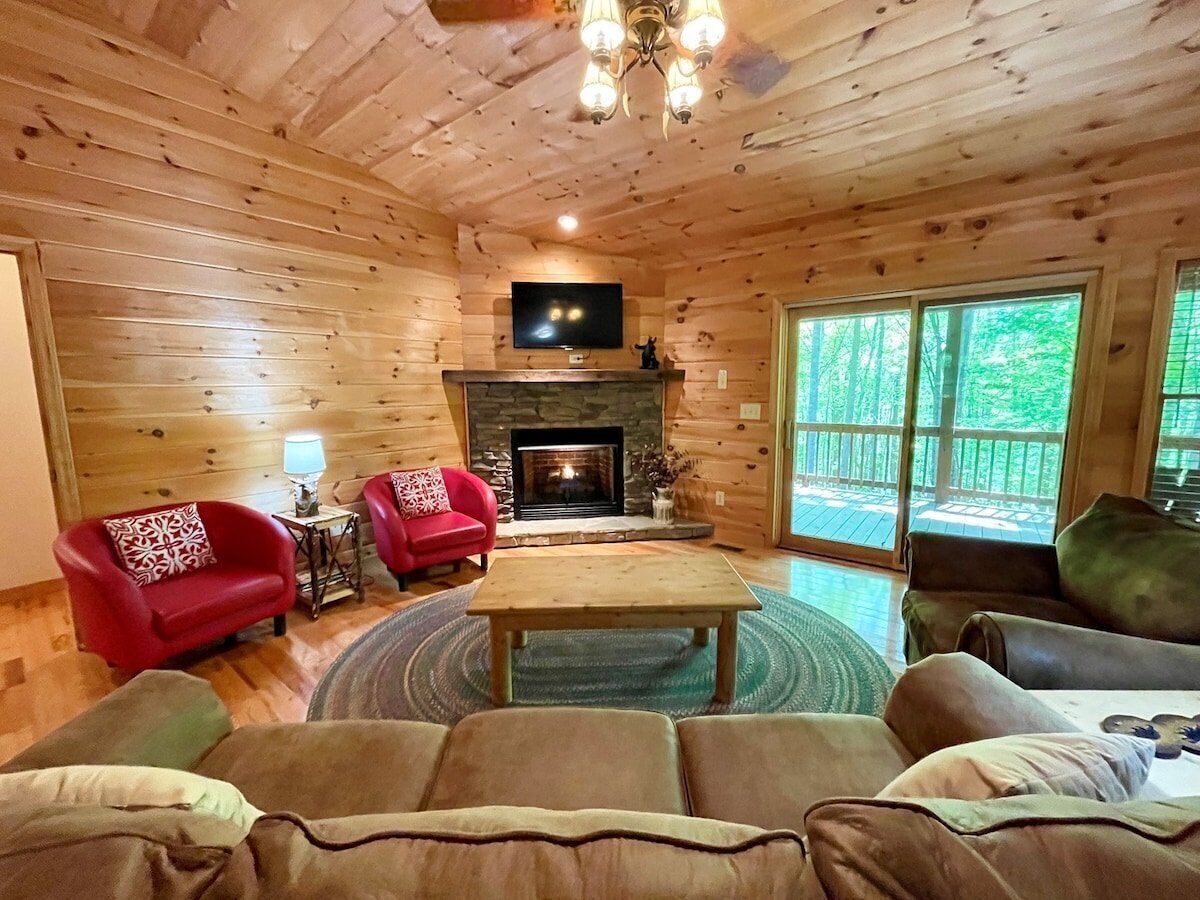 A cozy cabin interior with a fireplace, wooden walls, a television, and a large window.
