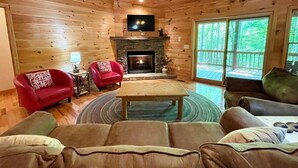 A cozy cabin interior with a fireplace, wooden walls, a television, and a large window.