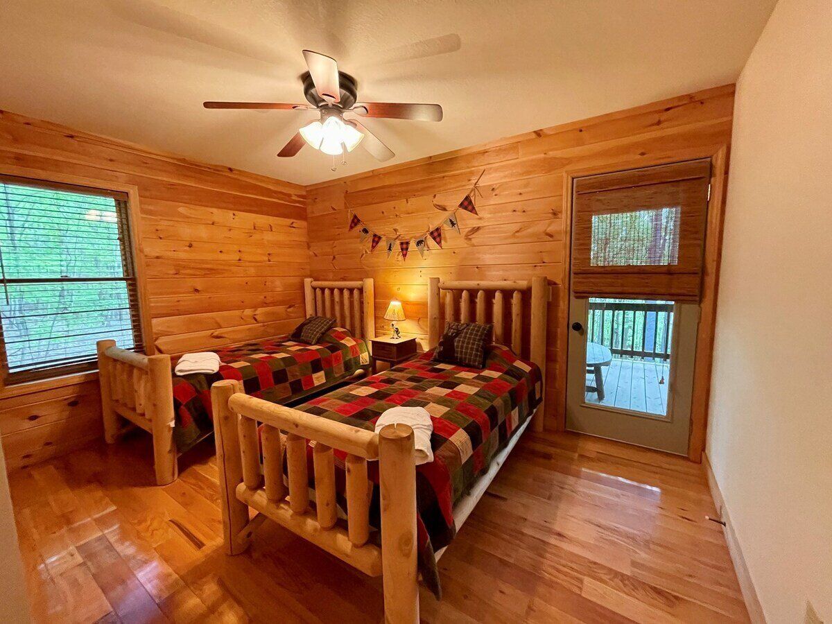 Un dormitorio tipo cabaña de madera con dos camas, un ventilador de techo y vistas al exterior a través de una puerta.