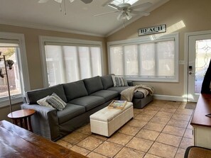 A living room with a large gray sectional sofa, a white ottoman, a small round table, and a ceiling fan.