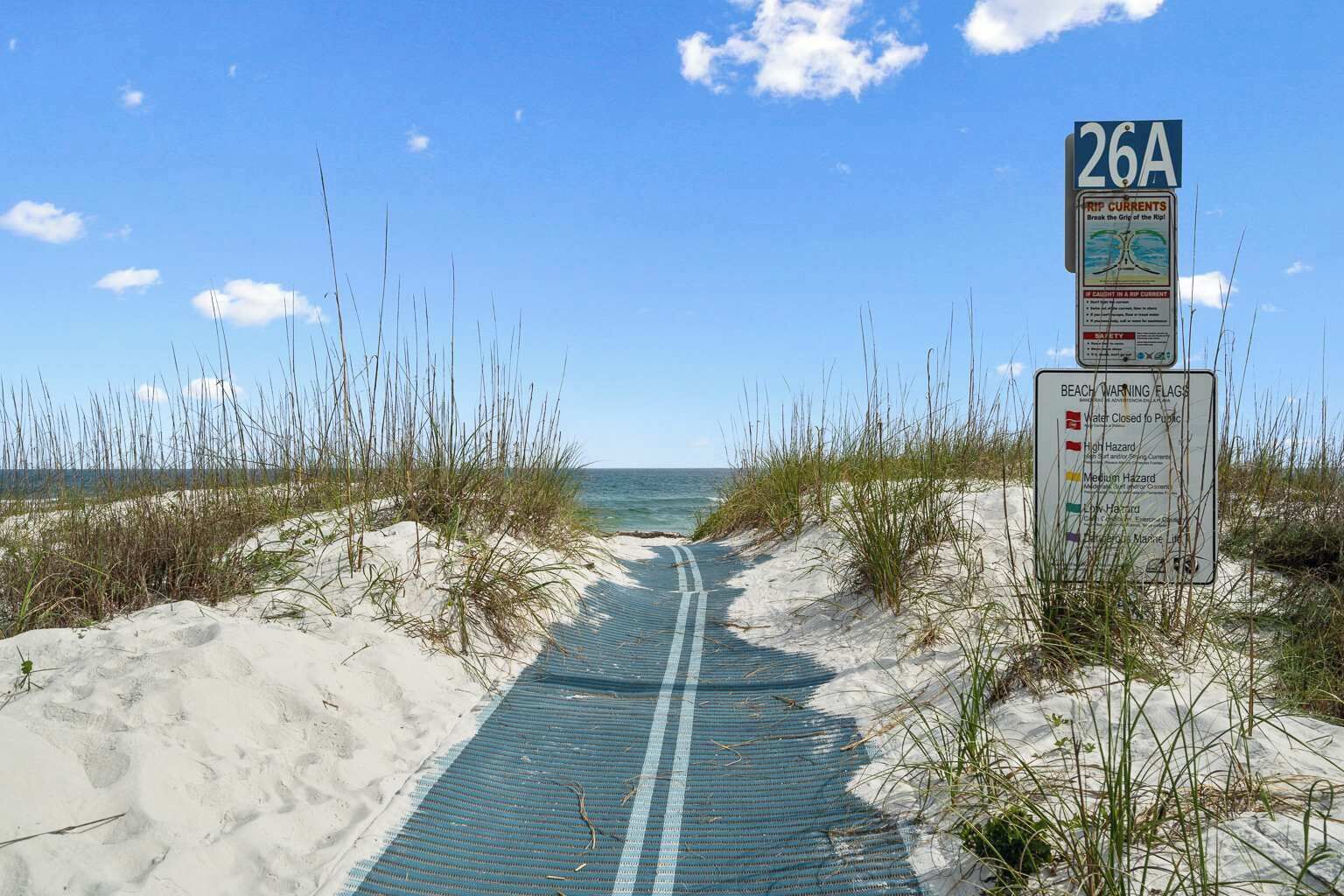 Un sentier qui longe la plage et mène à l’océan, avec des panneaux d’avertissement concernant les courants de contre-rivage et les dangers de la plage.