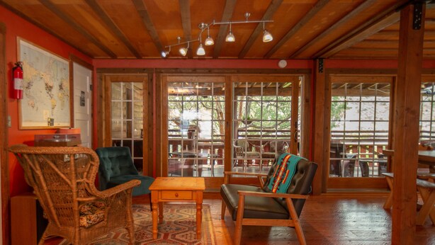 A cozy room with a large wooden ceiling, a wicker rocking chair, a teal armchair, and a dining table with chairs.
