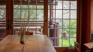 A wooden dining table with a clear view through a large window to an outdoor area with a wooden structure and greenery.