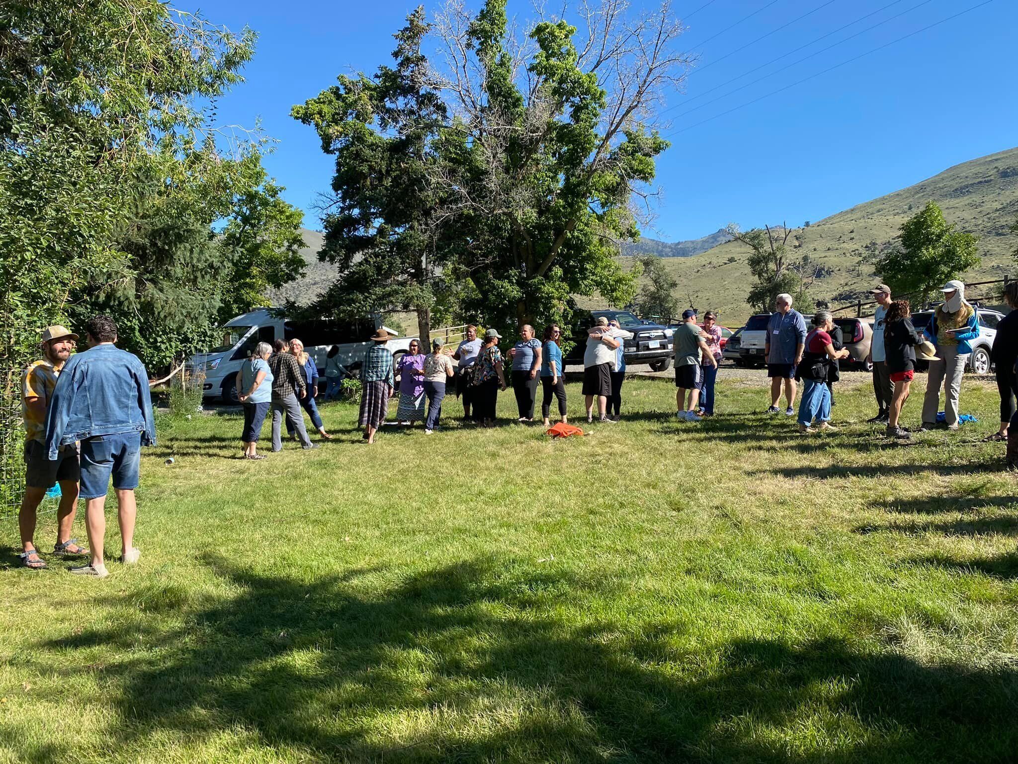 A group of people gathered outdoors in a grassy area with trees and hills in the background.