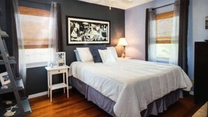 Un dormitorio con cama, mesita de noche, ventilador de techo y ventana con cortinas.
