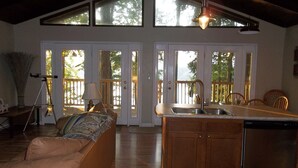 A spacious living area with a sofa, a dining table with chairs, a kitchen with a sink and dishwasher, and large windows with a view of trees. -  Lake Front Home with community boat ramp. Private,  close to marinas and town. (Somerset)