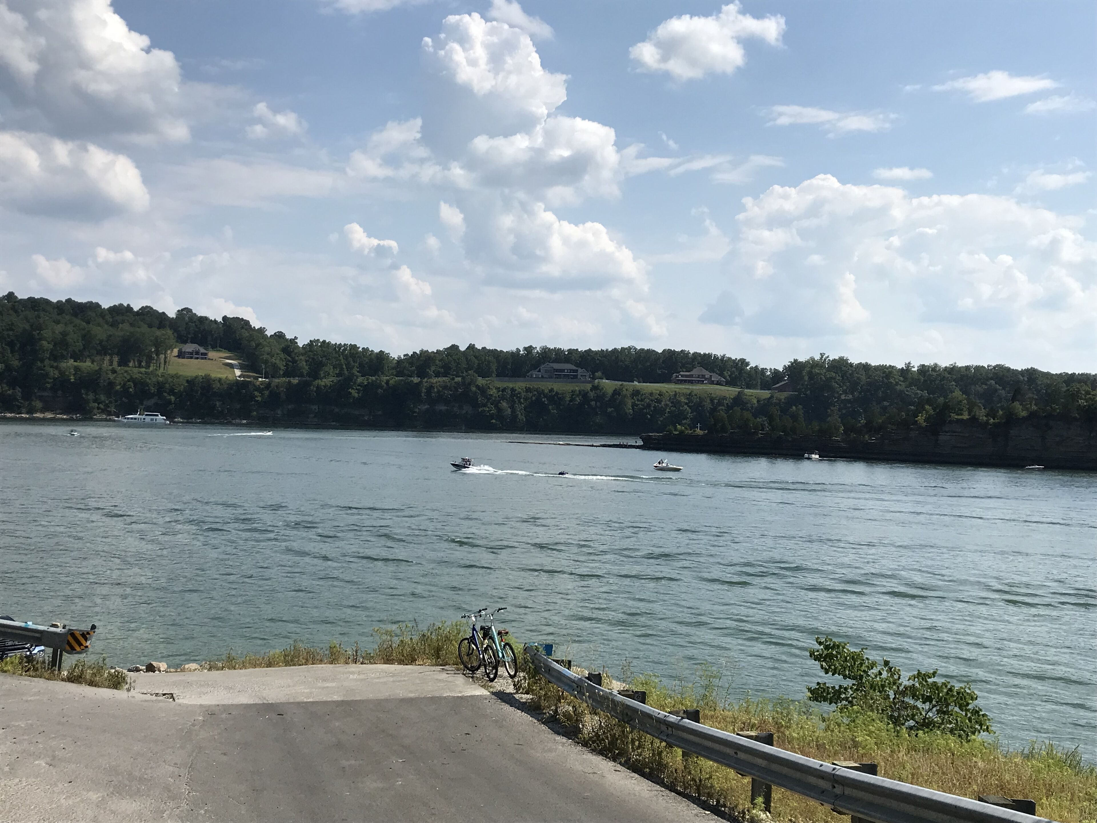 Vista de um lago com barcos, uma área pavimentada e um corrimão de metal.