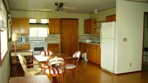 A compact kitchen with wooden cabinets, a dining table with chairs, a ceiling fan, and a refrigerator.