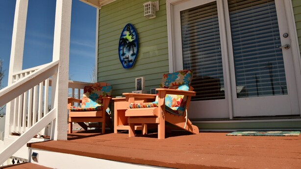 A porch with two wooden chairs and a table, a green house wall, and a window with blinds. - Bettie's Beach House (Mexico Beach)