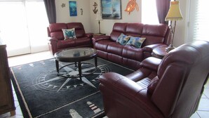 A living room with a red leather sofa, a coffee table, and a floor lamp. There is a painting on the wall and a window with curtains. - Bettie's Beach House (Mexico Beach)