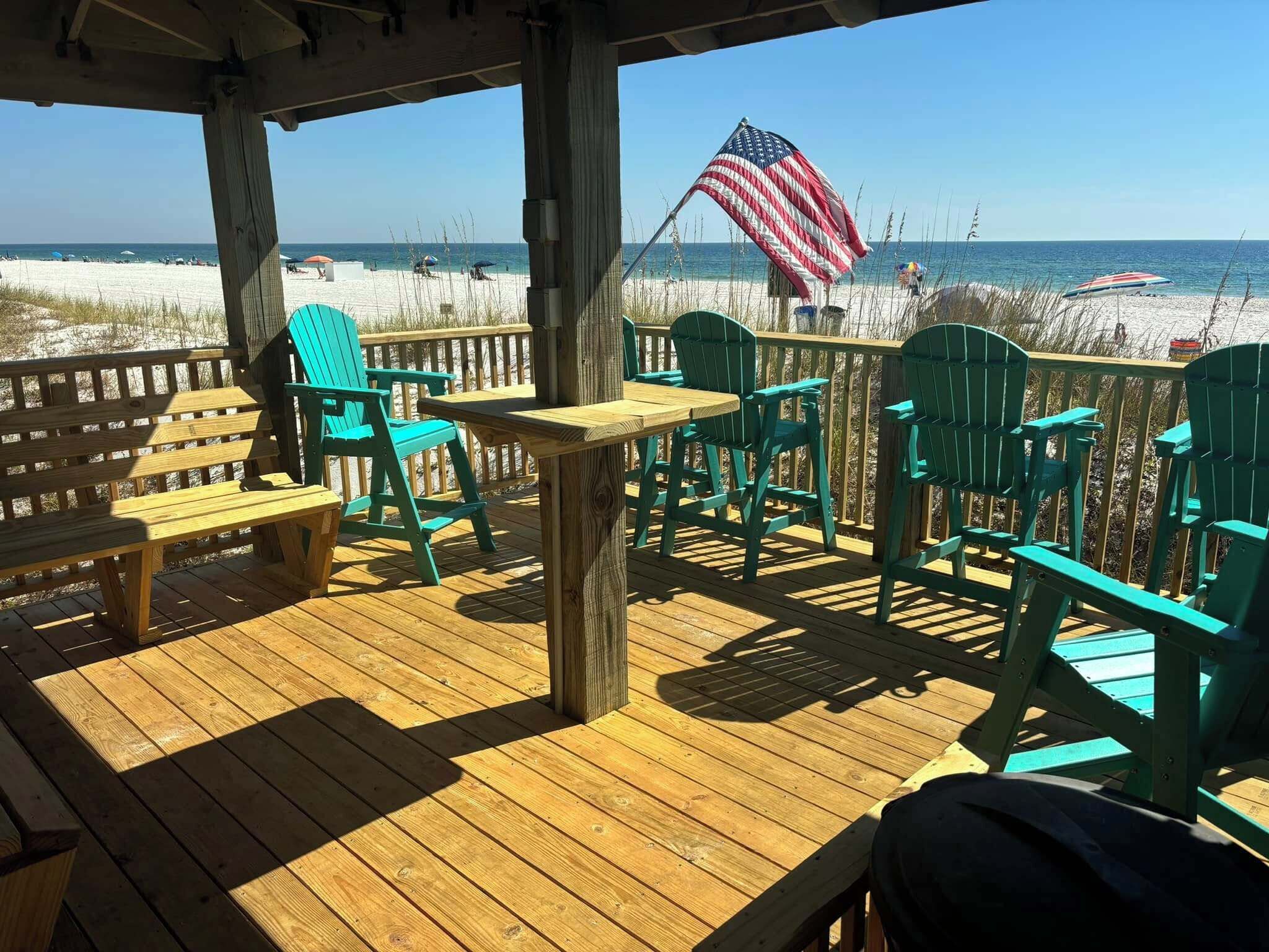 Um deck de madeira com cadeiras Adirondack verdes e uma mesa, com vista para uma praia e uma bandeira americana.