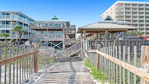 Een houten looppad leidt naar een strandhotel met meerdere balkons en een overdekt gedeelte.