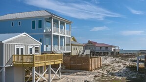 Une maison située en bord de mer, dotée d’un balcon à deux étages, d’une terrasse en bois et, en arrière-plan, d’une plage de sable.