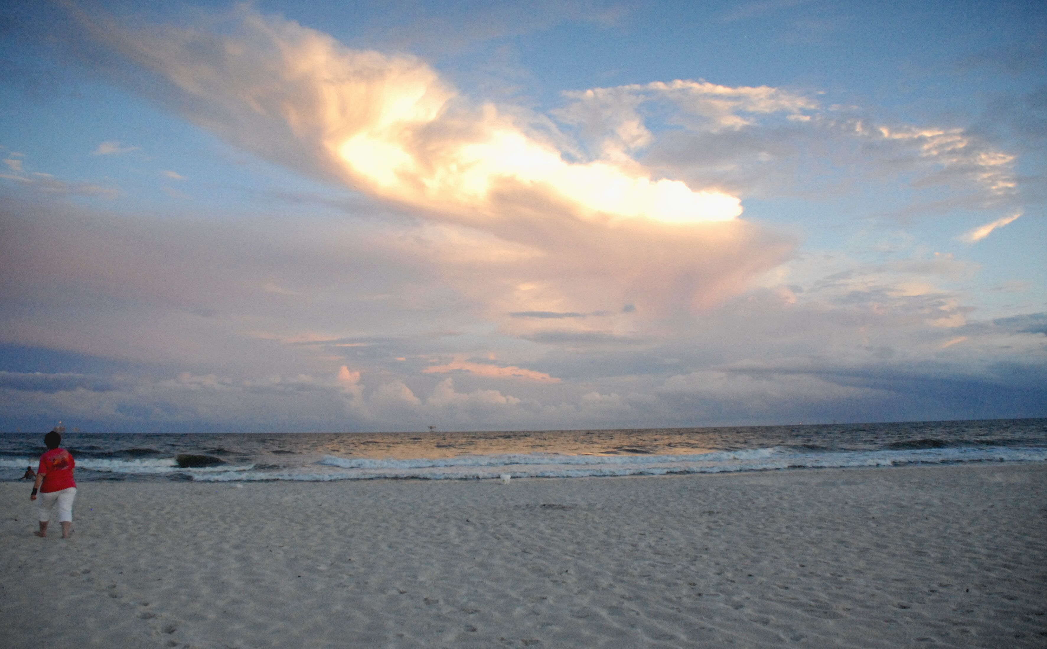 A beach with a person walking on the sand, waves approaching the shore, and a cloudy sky.