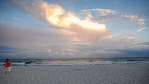Une plage où l’on voit une personne se promener sur le sable, des vagues qui se rapprochent du rivage et un ciel nuageux.