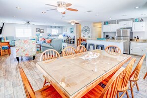Ein geräumiges Wohnzimmer mit großem Esstisch aus Holz, bequemen Sitzgelegenheiten und einer Küchenzeile mit weißen Schränken und modernen Geräten.