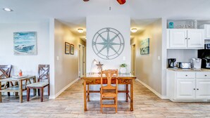 Un coin repas avec une table et des chaises en bois, une décoration murale représentant une boussole, et une cuisine avec des meubles blancs et une machine à café.
