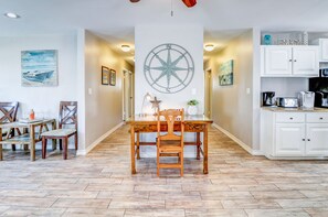 Un coin repas avec une table et des chaises en bois, une décoration murale représentant une boussole, et une cuisine avec des meubles blancs et une machine à café.