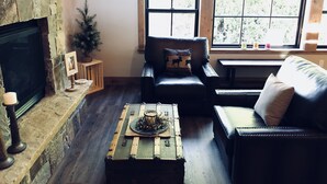 A cozy living room with a stone fireplace, leather furniture, and a view through large windows.