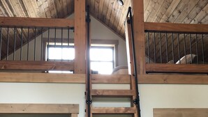 A loft bedroom with a wooden ladder leading to a lofted area, a bed with a patterned bedspread, and a window with curtains.